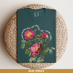 May include: A teal blue t-shirt with a floral design and the text "Your silence will not protect you. - Audre Lorde".