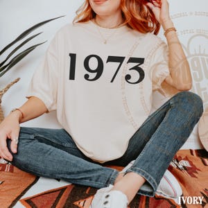 May include: A cream-colored short-sleeve t-shirt featuring the black text 1973 printed in a bold serif font across the chest. The shirt has a crew neckline and a loose, relaxed fit, paired with dark blue denim jeans and white sneakers.