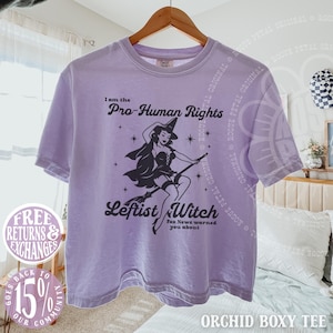May include: A light purple short-sleeve t-shirt featuring a black graphic of a witch on a broomstick. The text reads: I am the Pro-Human Rights Leftist Witch Fox News warned you about. The shirt has a crew neckline and a boxy, relaxed fit.