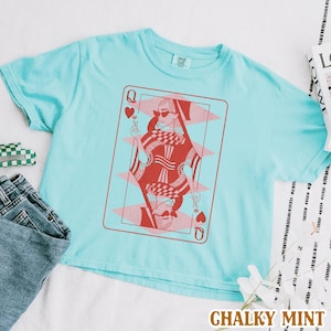 以下が含まれることがあります： 薄い青色のTシャツに、赤とピンクのハートの女王のトランプのグラフィックがプリントされています。グラフィックには、サングラスと王冠をつけた女性が描かれています。