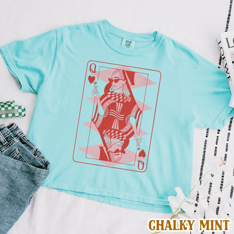 以下が含まれることがあります： 薄い青色のTシャツに、赤とピンクのハートの女王のトランプのグラフィックがプリントされています。グラフィックには、サングラスと王冠をつけた女性が描かれています。