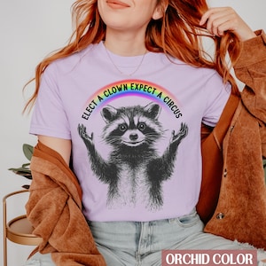May include: A purple t-shirt with a rainbow arc above a black and white raccoon. The text on the shirt reads "Elect a Clown Expect a Circus".