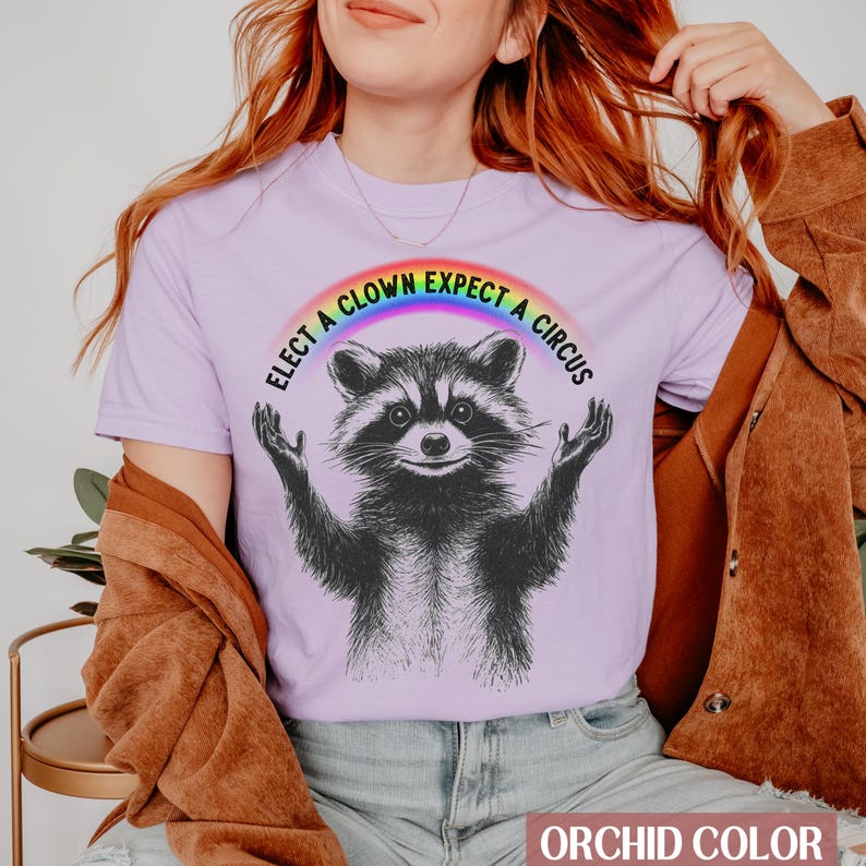May include: A purple t-shirt with a rainbow arc above a black and white raccoon. The text on the shirt reads "Elect a Clown Expect a Circus".