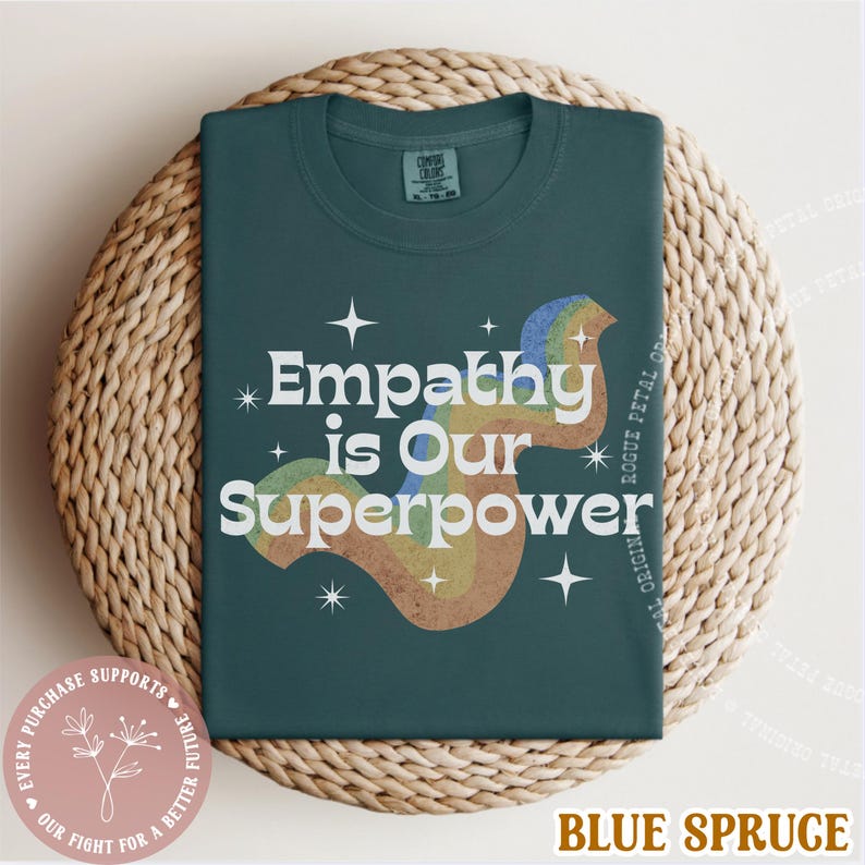 Puede incluir: Camiseta azul abeto con el texto "Empathy is Our Superpower" en blanco, con un dise&ntilde;o de arco&iacute;ris. La camiseta est&aacute; doblada sobre una cesta tejida. El texto adicional incluye "Cada compra apoya nuestra lucha por un futuro mejor."