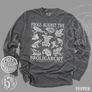 May include: Pepper-colored long-sleeve shirt with a white graphic of various frog illustrations. The text "FROGS AGAINST THE BROLIGARCHY THE RIBBOLUTION IS NOW" is printed on the front. The shirt has a crew neck and long sleeves.