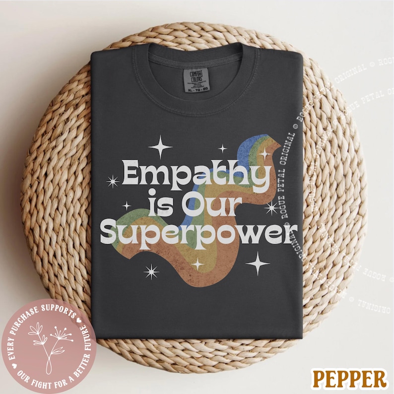 Puede incluir: Una camiseta gris oscuro con el texto "Empathy is Our Superpower" en blanco, con un dise&ntilde;o de arco&iacute;ris. La camiseta se muestra sobre una superficie circular tejida. La palabra "Pepper" est&aacute; en la parte inferior derecha.