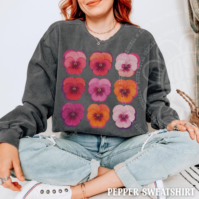 Puede incluir: Una sudadera de cuello redondo en color gris carb&oacute;n con una cuadr&iacute;cula de nueve ilustraciones de flores de pensamiento en tonos rosa, morado y naranja. La prenda tiene mangas largas, cuello acanalado y una textura de tela lavada vintage.