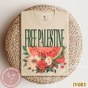 May include: Ivory-colored t-shirt with the text "FREE PALESTINE" in green. The design includes a watermelon slice and floral elements in red, pink, and white. The shirt is folded on a woven mat.