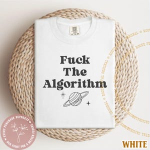 May include: White t-shirt with the text "Fuck The Algorithm" in a bold, black font. A graphic of a planet with a ring and a star are below the text. The shirt is folded on a woven, circular surface.