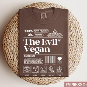 May include: Espresso brown t-shirt with a white graphic that reads "100% Plant-Powered 0% Regrets The Evil* Vegan" with a barcode and icons representing animal lover, planet activist, gourmet foodie, half human half tofu, and vegan spot GPS.