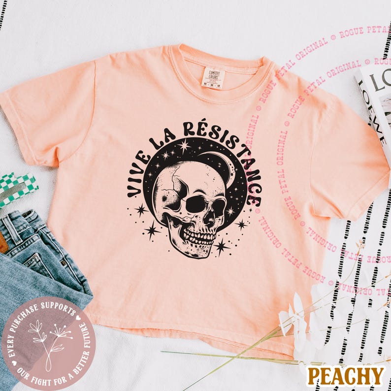 May include: Peach-colored crop top with a black and white skull graphic. The text "VIVE LA R&Eacute;SISTANCE" is above the skull. The shirt is made of a soft material and is likely part of a casual wear collection.