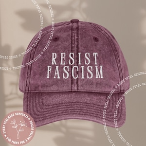 May include: A burgundy baseball cap with the embroidered text "RESIST FASCISM" in white. The hat has a curved brim and a button on top. The design includes a circular graphic with text and floral elements.
