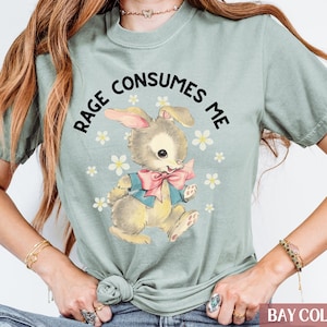 Op de afbeelding: Een lichtgroen T-shirt met een cartoonkonijn dat een blauwe en roze strik draagt. De tekst "RAGE CONSUMES ME" is in zwart boven het konijn gedrukt.