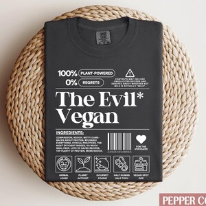 May include: Black t-shirt with white text that reads "100% Plant-Powered 0% Regrets The Evil* Vegan". The shirt has a graphic of a barcode, a heart with the text "For the Voiceless", and a series of icons representing different aspects of veganism.