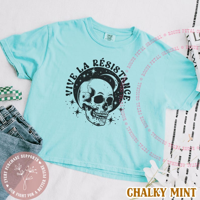 May include: A light blue t-shirt with the words "VIVE LA R&Eacute;SISTANCE" and a skull graphic. The shirt has short sleeves and a crew neck. The text "ROGUE PETAL ORIGINAL" is printed around the shirt.
