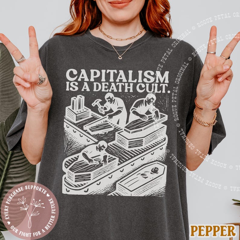 K&ouml;nnte beinhalten: Dunkelgraues T-Shirt mit dem Text "CAPITALISM IS A DEATH CULT." Die Grafik zeigt Arbeiter, die S&auml;rge an einem Flie&szlig;band bauen. Ein rundes Logo mit Text und floralem Design ist unten links sichtbar.