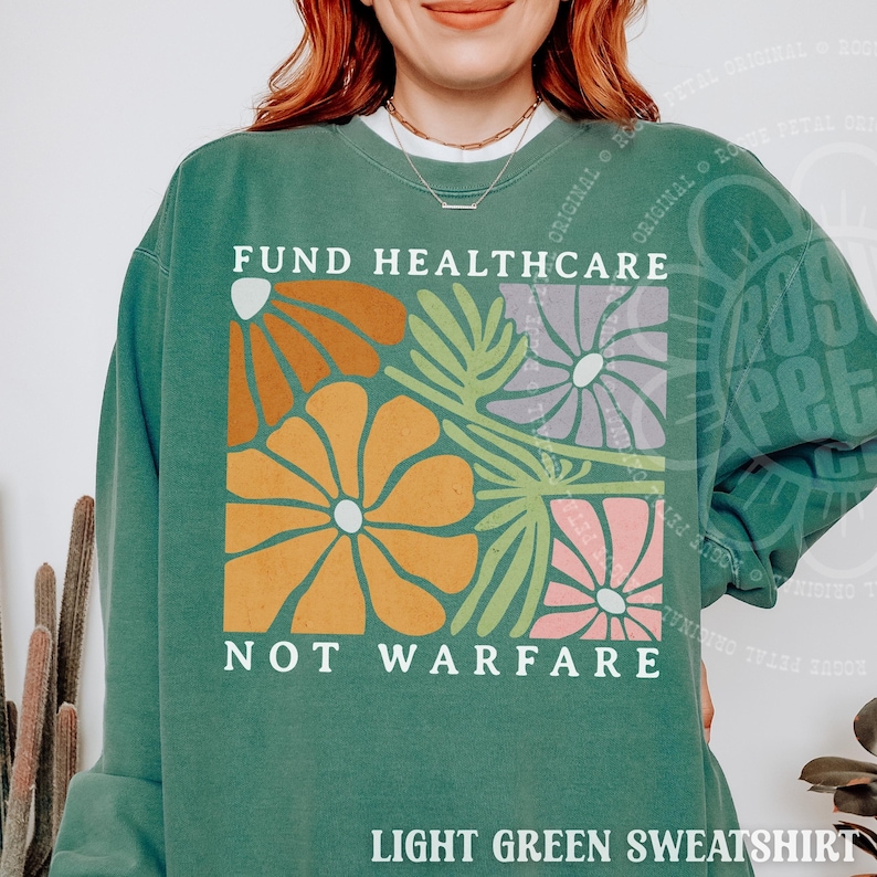 May include: A light green crewneck sweatshirt featuring a graphic print of stylized flowers in orange, yellow, lavender, and pink. The text "FUND HEALTHCARE NOT WARFARE" is printed in white capital letters above and below the floral design.