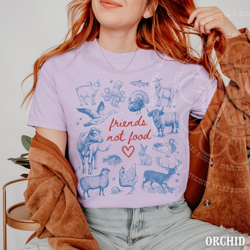Puede incluir: Camiseta lila claro con una ilustraci&oacute;n azul de varios animales. El texto "friends not food" est&aacute; escrito en rojo con un coraz&oacute;n. La camiseta tiene cuello redondo y est&aacute; hecha de un material suave.