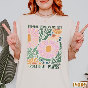 May include: A cream-colored t-shirt with a floral design and the text "Federal Workers Are Not Political Pawns".