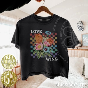 May include: Black boxy tee shirt with a floral bouquet graphic. The shirt features the words "LOVE WINS" in white text. The floral design includes orange, pink, and blue flowers against a checkered background.