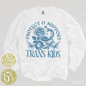 May include: White long-sleeve shirt with a blue graphic. The graphic includes a rose, snake, butterflies, and text that reads "PROTECT & SUPPORT TRANS KIDS." Additional text includes "FREE RETURNS & EXCHANGES" and "GOES BACK TO 15% OUR COMMUNITY."