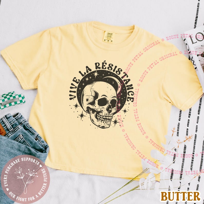 May include: Butter-colored t-shirt featuring a black and white skull design with the words "VIVE LA R&Eacute;SISTANCE" in a circular pattern. The shirt has short sleeves and a crew neck. The text "ROGUE PETAL ORIGINAL" is printed around the shirt.