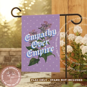 May include: A purple garden flag with white polka dots and the words "Empathy Over Empire" in a decorative font. The flag features a floral illustration and is displayed on a black metal stand. The flag is approximately 12 inches wide.