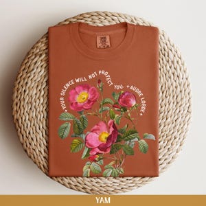 May include: A rust-colored t-shirt with a floral design and the text "Your silence will not protect you. - Audre Lorde".