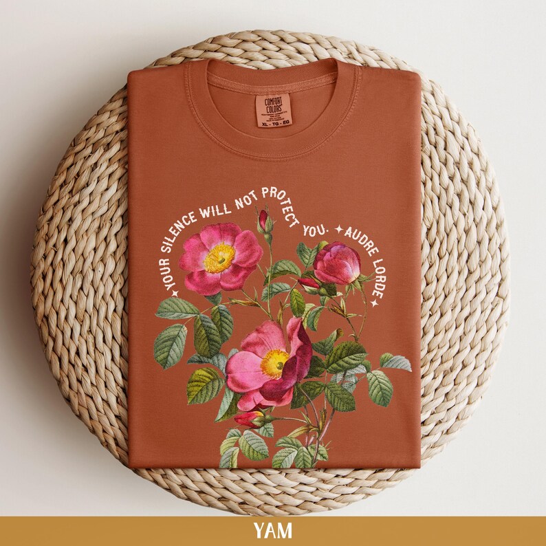 May include: A rust-colored t-shirt with a floral design and the text "Your silence will not protect you. - Audre Lorde".