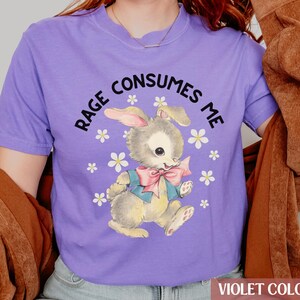 Op de afbeelding: Een paars T-shirt met een afbeelding van een cartoonkonijn met de tekst "RAGE CONSUMES ME" erop gedrukt.