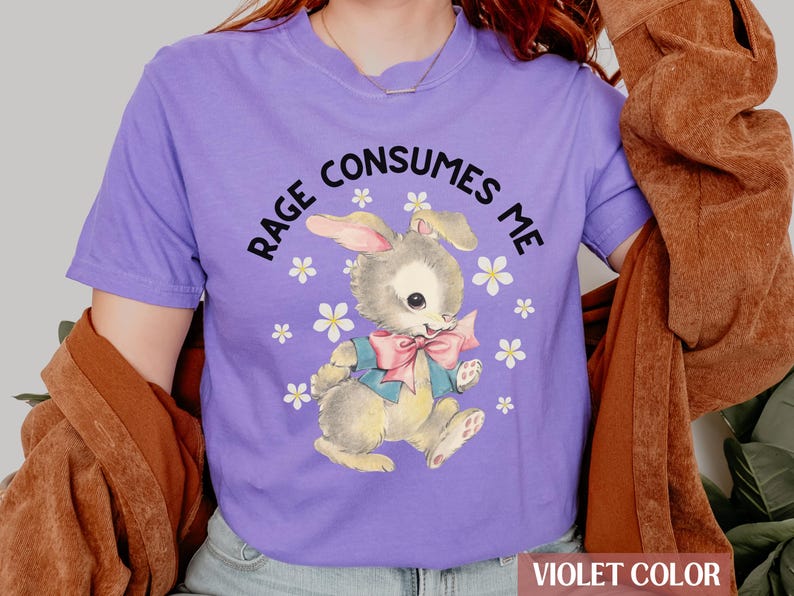 Op de afbeelding: Een paars T-shirt met een afbeelding van een cartoonkonijn met de tekst "RAGE CONSUMES ME" erop gedrukt.