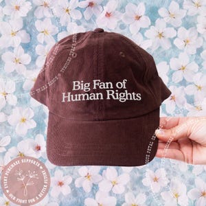 May include: A brown corduroy baseball cap with the embroidered text "Big Fan of Human Rights" in white. The cap has a curved brim and a rounded top. The background features a floral pattern.