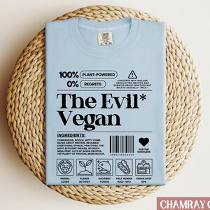 May include: Light blue t-shirt with a white graphic design that says "100% Plant-Powered 0% Regrets The Evil* Vegan" with a barcode and icons below the text. The icons represent animal lover, planet activist, gourmet foodie, half human half tofu, and vegan spot GPS.
