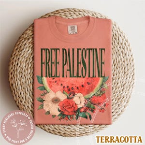 May include: Terracotta-colored t-shirt with the text "FREE PALESTINE" in dark green. The shirt features a watermelon slice and floral design. The shirt is folded on a woven, circular surface.