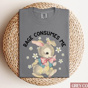 Op de afbeelding: Een grijs T-shirt met de tekst "Rage Consumes Me" en een cartoonkonijn met een blauwe en roze strik. Het konijn is omringd door witte bloemen.