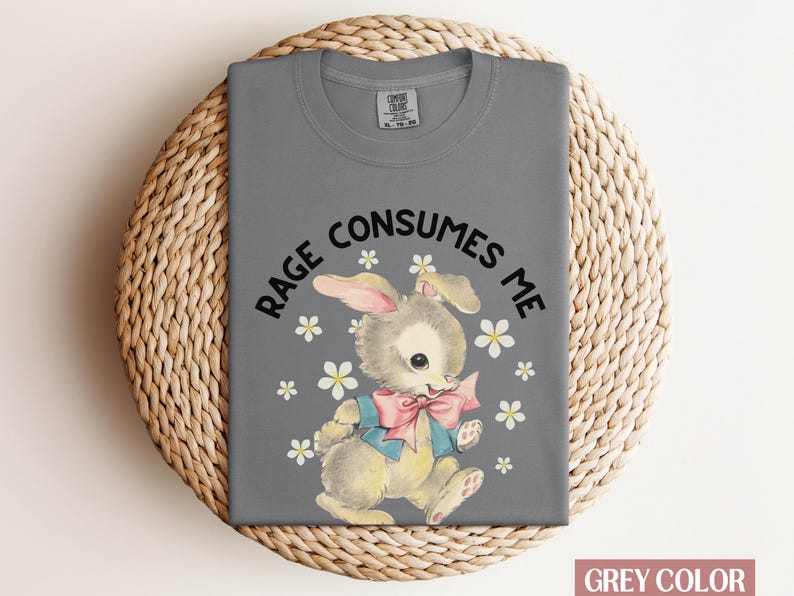Op de afbeelding: Een grijs T-shirt met de tekst "Rage Consumes Me" en een cartoonkonijn met een blauwe en roze strik. Het konijn is omringd door witte bloemen.