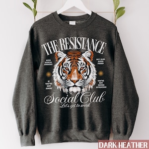 May include: Dark heather grey sweatshirt with a graphic print of a tiger and the text "THE RESISTANCE Social Club Let's get to work". The graphic includes the text "With radical empathy" and "But also super pissed".