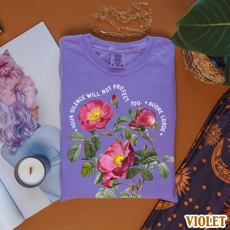 May include: Purple t-shirt with a floral design and the text "Your silence will not protect you. Audre Lorde".
