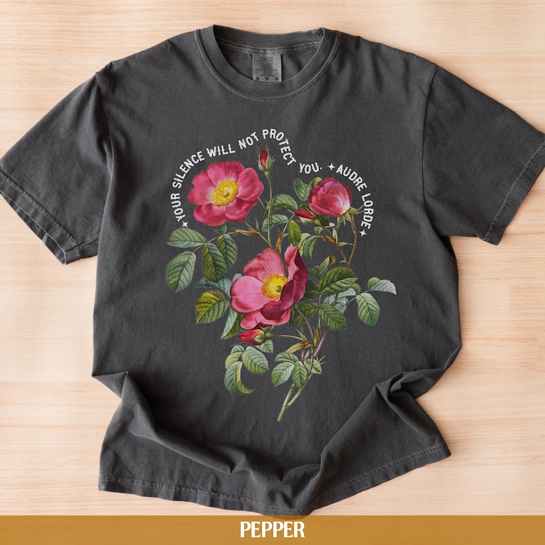 May include: A dark gray t-shirt with a floral design and the text "Your silence will not protect you. - Audre Lorde" printed in white.