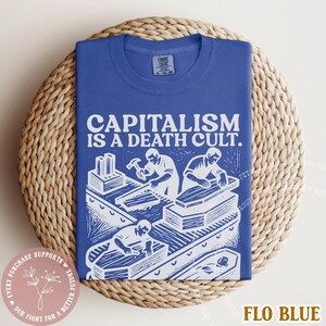 K&ouml;nnte beinhalten: K&ouml;nigsblaues T-Shirt mit dem Text "CAPITALISM IS A DEATH CULT." und einer wei&szlig;en Illustration von Arbeitern, die S&auml;rge bauen. Das T-Shirt liegt gefaltet auf einem runden Korb.