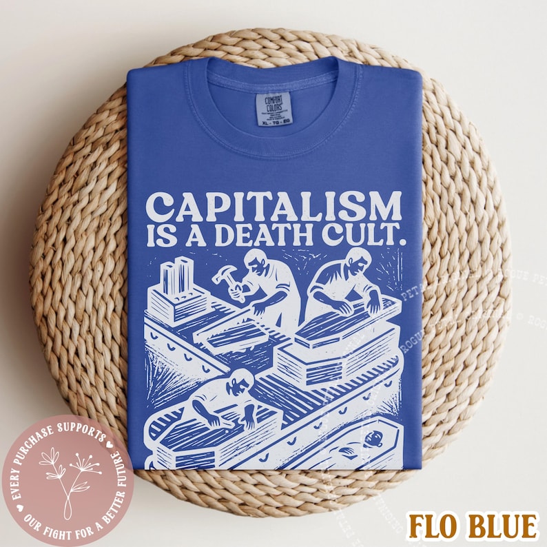K&ouml;nnte beinhalten: K&ouml;nigsblaues T-Shirt mit dem Text "CAPITALISM IS A DEATH CULT." und einer wei&szlig;en Illustration von Arbeitern, die S&auml;rge bauen. Das T-Shirt liegt gefaltet auf einem runden Korb.