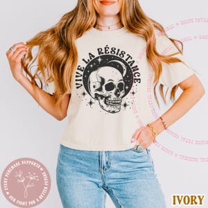 May include: Ivory-colored t-shirt featuring a black and white skull design with the words "VIVE LA R&Eacute;SISTANCE" in a circular pattern. The shirt also has star and moon graphics. The text "IVORY" is at the bottom.
