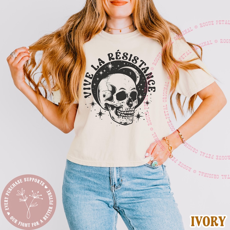 May include: Ivory-colored t-shirt featuring a black and white skull design with the words "VIVE LA R&Eacute;SISTANCE" in a circular pattern. The shirt also has star and moon graphics. The text "IVORY" is at the bottom.