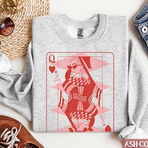 May include: A gray sweatshirt with a red queen of hearts playing card graphic. The queen is wearing sunglasses and a crown. The text "ASH COLOR" is visible at the bottom of the image.