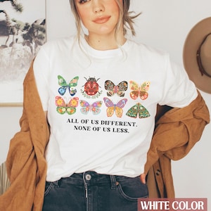May include: White t-shirt with a colorful butterfly and ladybug design. The text "All of us different. None of us less." is printed on the shirt.