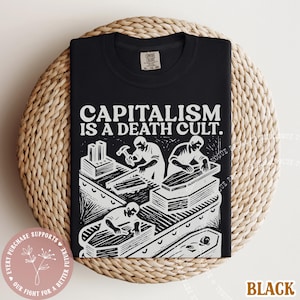 K&ouml;nnte beinhalten: Schwarzes T-Shirt mit dem Text "CAPITALISM IS A DEATH CULT." und einer wei&szlig;en Grafik von Arbeitern, die S&auml;rge bauen. Das Shirt liegt auf einer runden, gewebten Unterlage.