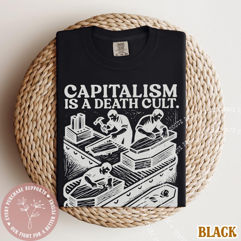 K&ouml;nnte beinhalten: Schwarzes T-Shirt mit dem Text "CAPITALISM IS A DEATH CULT." und einer wei&szlig;en Grafik von Arbeitern, die S&auml;rge bauen. Das Shirt liegt auf einer runden, gewebten Unterlage.