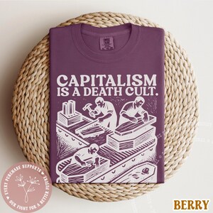 K&ouml;nnte beinhalten: Beerenfarbenes T-Shirt mit dem wei&szlig;en Text "CAPITALISM IS A DEATH CULT". Die Grafik zeigt Arbeiter und S&auml;rge. Das T-Shirt wird auf einer gewebten Oberfl&auml;che pr&auml;sentiert.