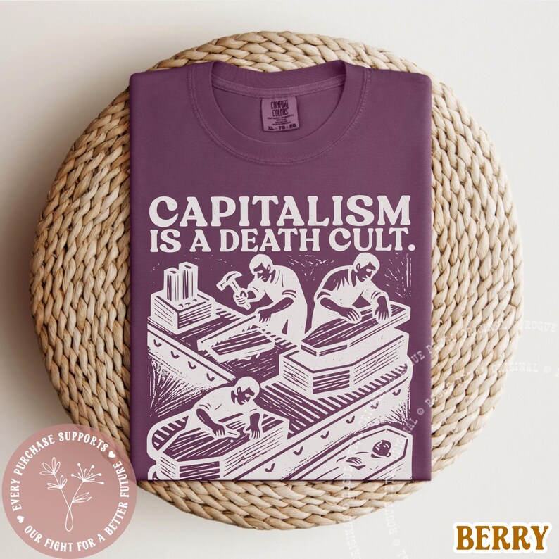 K&ouml;nnte beinhalten: Beerenfarbenes T-Shirt mit dem wei&szlig;en Text "CAPITALISM IS A DEATH CULT". Die Grafik zeigt Arbeiter und S&auml;rge. Das T-Shirt wird auf einer gewebten Oberfl&auml;che pr&auml;sentiert.