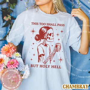 May include: Light blue t-shirt with a red skeleton graphic holding a coffee cup. The text on the shirt reads "THIS TOO SHALL PASS BUT HOLY HELL". The shirt is made of chambray material.